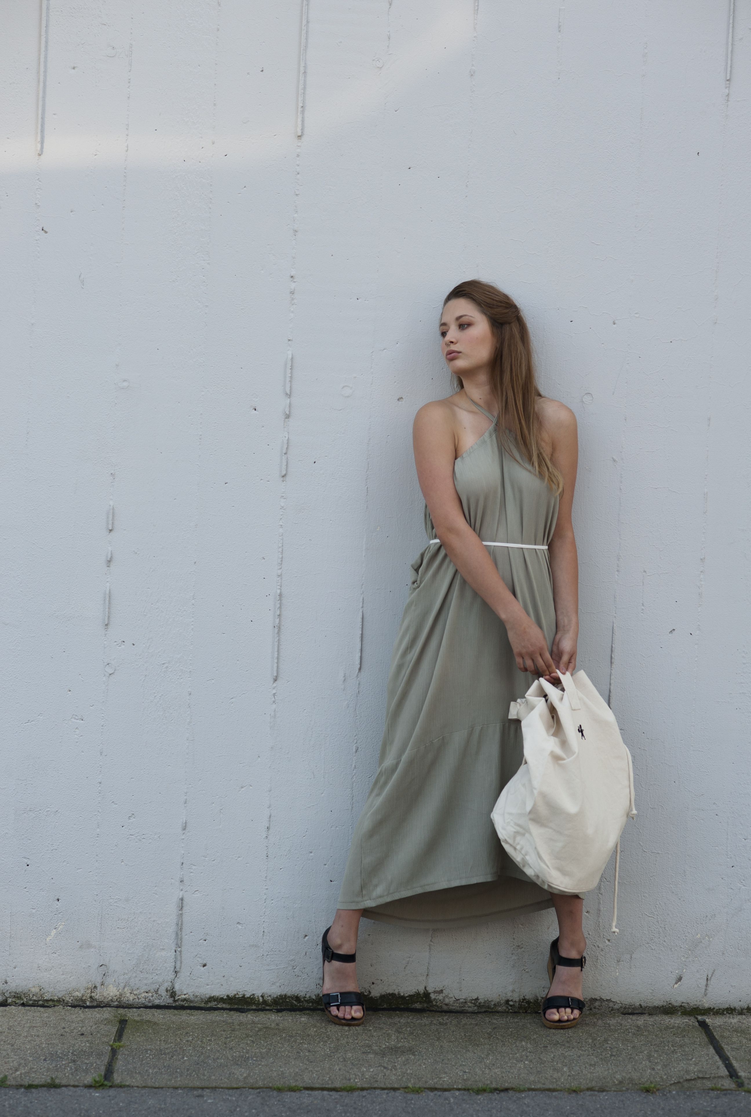 A girl holding a white backpack and wearing pale green long sleeveless fress tied behind her neck