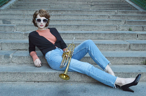 Model is lying on the stairs wearing light blue jeans, black bolero, orange turtle neck pullover and sunglasses with blue rim. She is holding golden trumpet