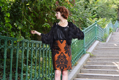 Woman standing on the stairs is wearing orange high waisted pencil skirt with baroque pattern and black vintage style top