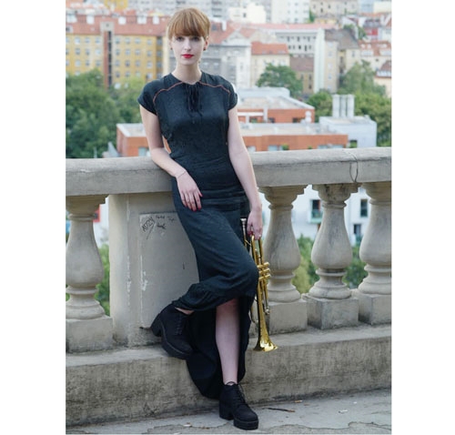 The model is wearing long blac drerss and she is holding golden a trumpet. There is a view on the city behind her