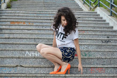 Woman is wearing recycled blue shorts with artistic yellow pattern, white T-shirt with black title WTF and orange high heels