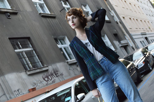 Woman standing in front of a building is wearing tartan jacket with green and blue stripes, black sleeves, pale blue jeans and white top